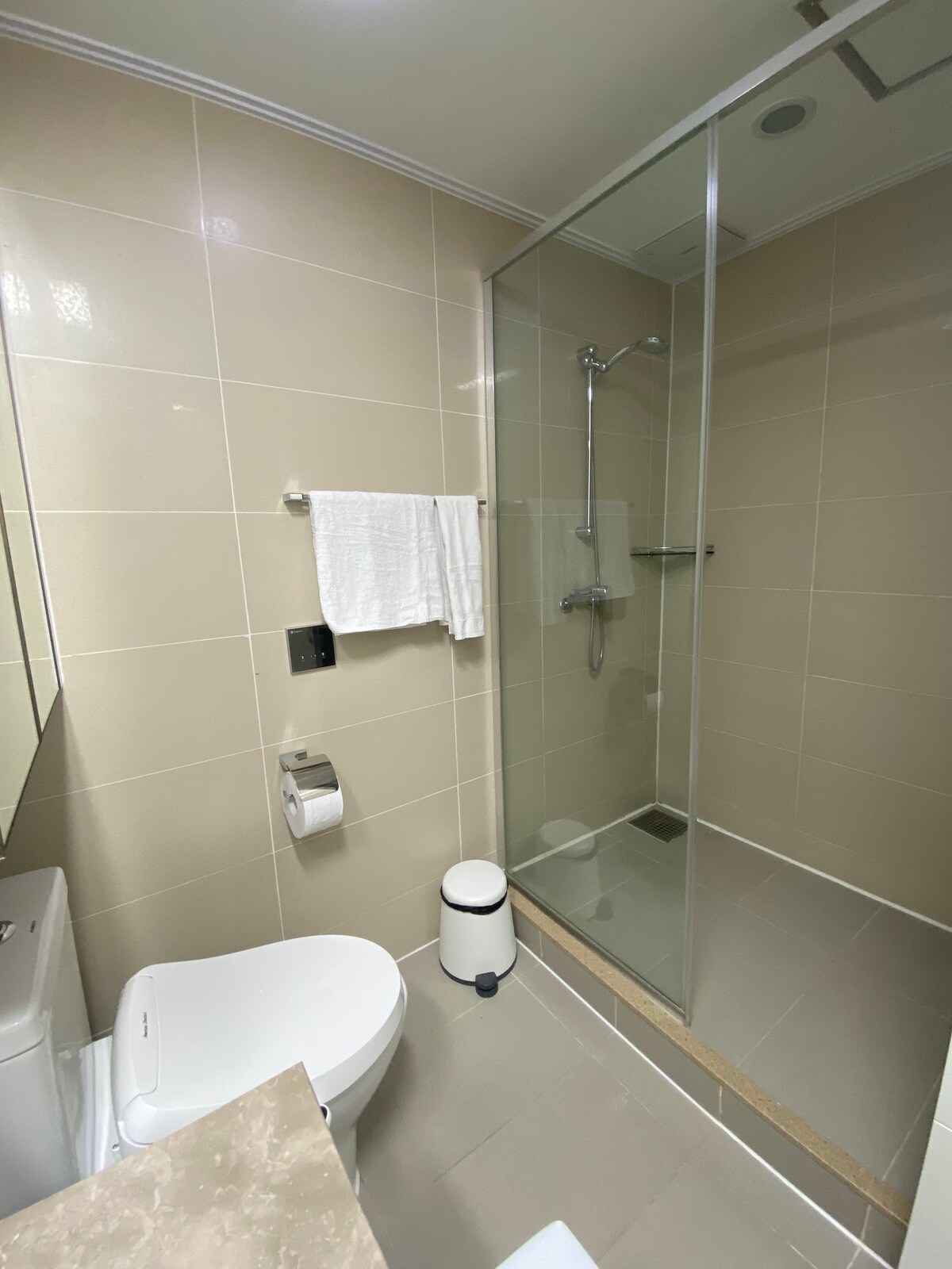 A modern bathroom features neutral tiles and sleek finishes. A glass shower enclosure is positioned beside the toilet. Soft white towels are neatly displayed on a rack. A small trash bin is placed on the floor, maintaining the room's clean aesthetic.