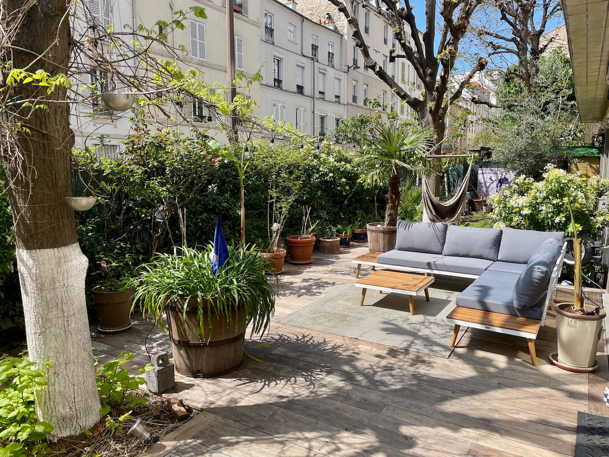 A private garden space features a comfortable seating arrangement with a sleek grey sofa and wooden side tables. Various potted plants surround the area, and a hammock is visible among the greenery. The garden setting provides a relaxing environment with ample natural light.