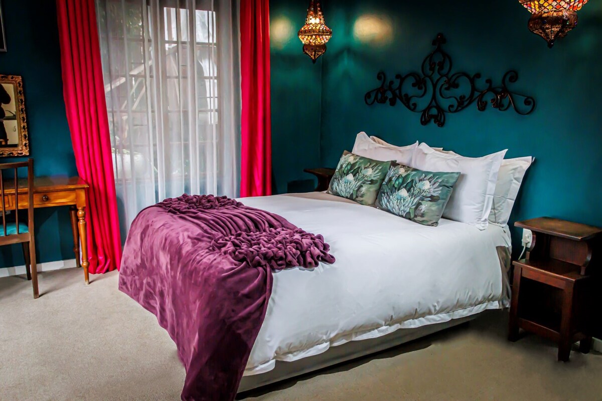 A cozy bedroom features a spacious bed dressed in crisp white linens and a deep plum throw. Green floral cushions sit atop the bed. Vibrant teal walls are complemented by pink curtains, while ornamental light fixtures add character to the space.