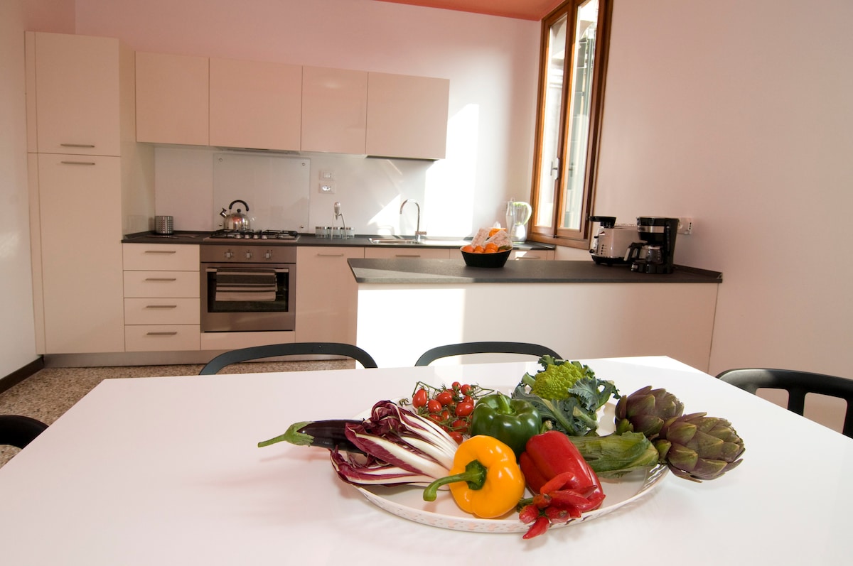 The kitchen features modern cabinetry and an array of appliances, including a stove and coffee maker. A dining table is set with a platter of fresh vegetables, showcasing a variety of colors and textures. Natural light floods the space through nearby windows.