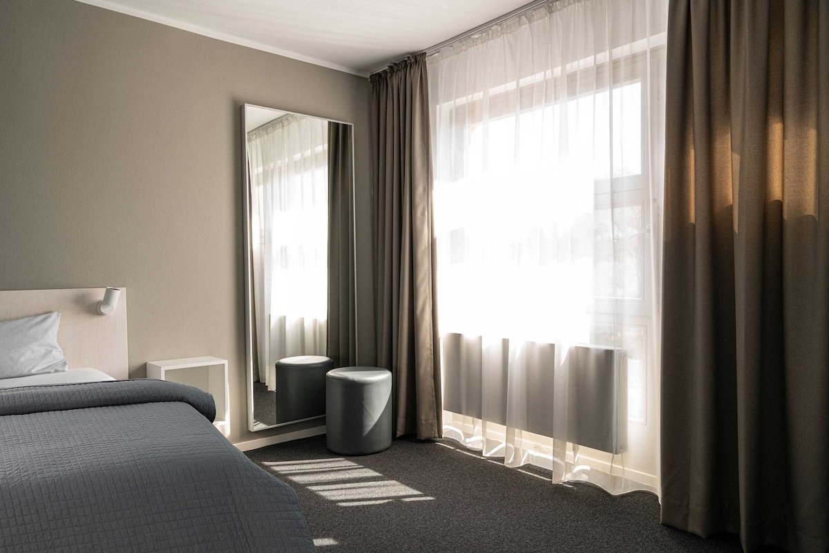 A well-lit room features soft natural light filtering through sheer curtains. A spacious bed is positioned near a large window, while a full-length mirror leans against the wall. A round stool complements the minimalist decor, contributing to the overall tranquil atmosphere.