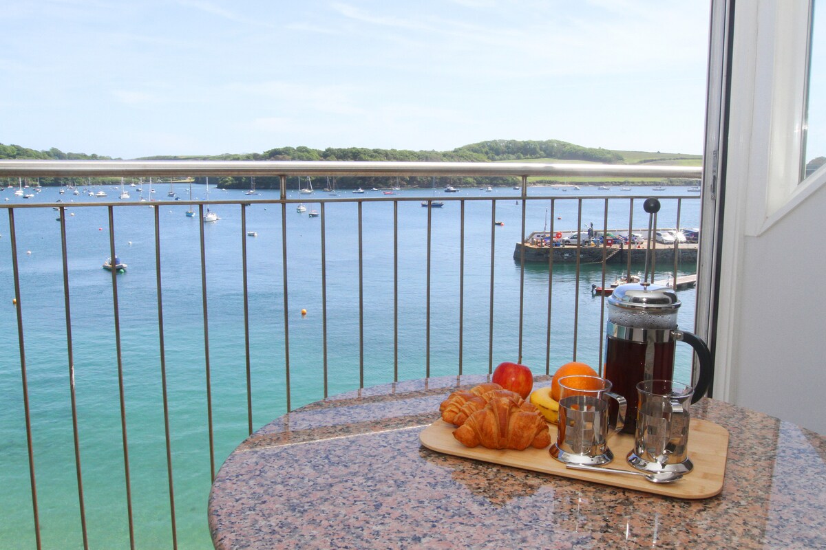 A balcony table is set with a wooden tray featuring croissants, fruit, and two glasses, accompanied by a French press. The unobstructed view showcases tranquil waters with anchored boats, framed by lush green hills under a clear blue sky.