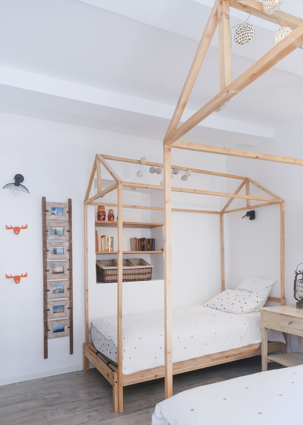 A bedroom features two unique beds designed in the shape of a house, presenting a playful atmosphere. A light, neutral palette is complemented by decorative elements on the walls. Shelving displays books and decorative items, providing a cozy and organized environment.