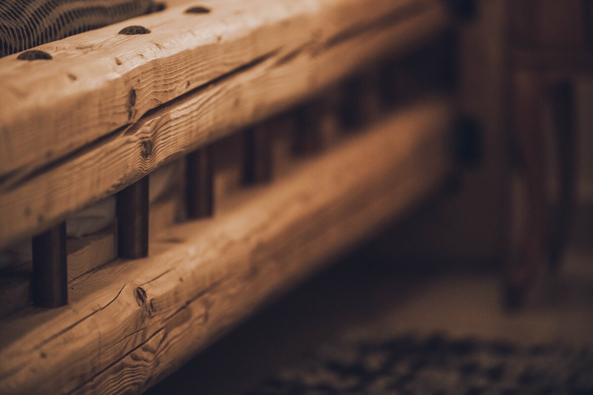 A close-up view of a rustic bed frame crafted from natural wood. The sturdy design features visible grains and textures, with wooden slats providing support underneath. A soft rug can be seen partially in the foreground, enhancing the overall warmth of the space.