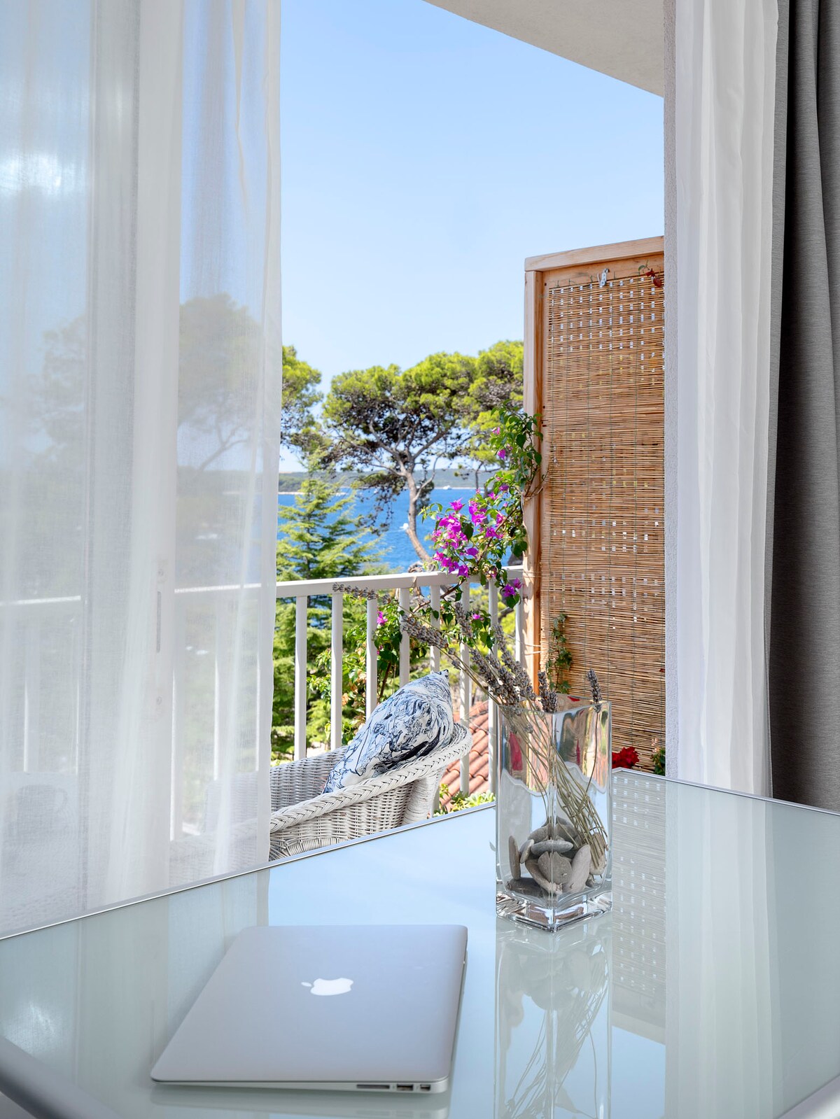 A serene balcony view is captured, featuring a small glass table with a laptop. Nearby, a floral arrangement adds a touch of nature. Soft curtains gently frame the scene, which overlooks lush greenery and a glimpse of the sea beyond.