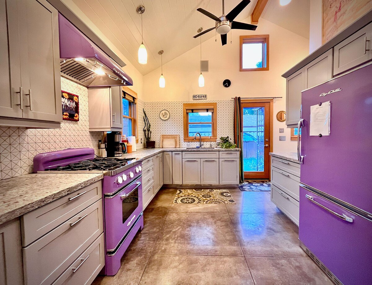 An artisan kitchen is highlighted by modern amenities and vibrant purple appliances. Ample storage is provided by stylish cabinetry, while the countertops feature a light stone finish. Natural light fills the room through a window above the sink, enhancing the warm atmosphere.