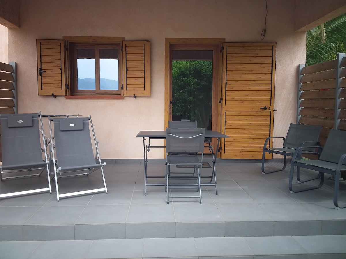 A shaded outdoor patio area is presented with a rectangular table and several armchairs. Two reclining lounge chairs are positioned to one side. Wooden shutters frame the windows, adding a natural element to the space, while potted greenery offers a touch of nature.