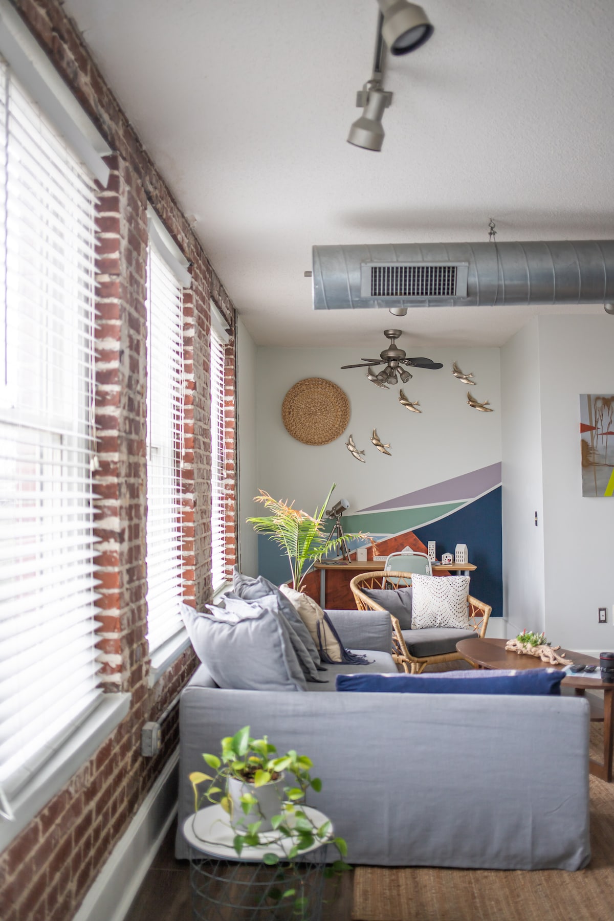 A living area features exposed brick walls and large windows adorned with blinds, allowing natural light to enter. A comfortable sofa is complemented by a mix of chairs and decorative elements, including wall art and a ceiling fan. Greenery is positioned on a side table for added freshness.
