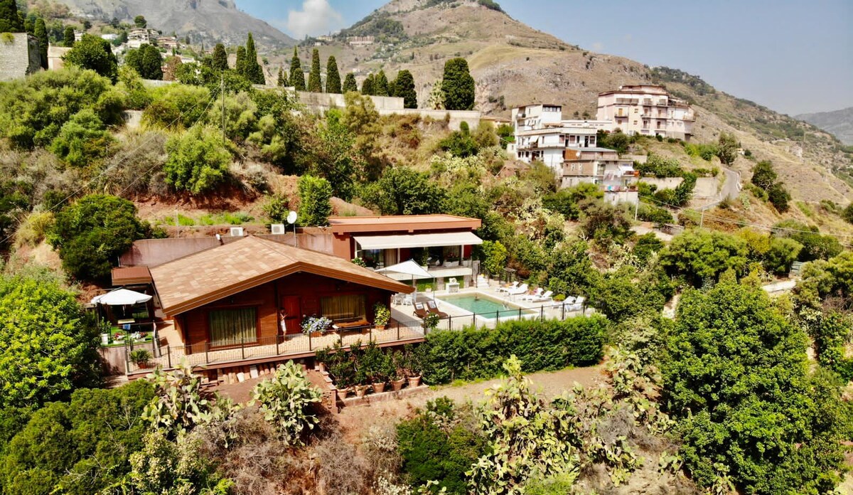 The villa is positioned on a hillside, surrounded by lush greenery and trees. The layout includes multiple buildings, with a solar-heated swimming pool visible in the shared area. Views of the surrounding mountains and valleys are prominent under a clear sky.