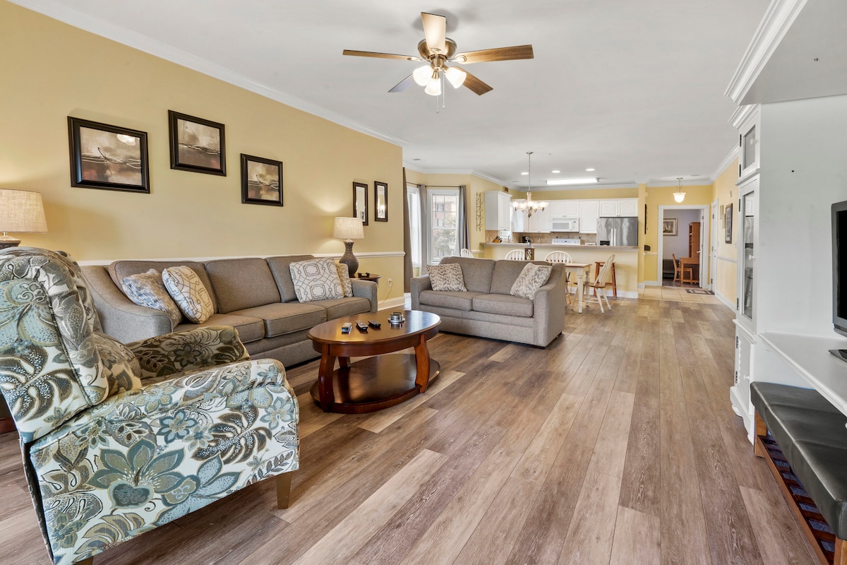 A spacious living area is furnished with two comfortable sofas and an armchair featuring a floral pattern. A coffee table sits centrally, and artwork adorns the walls. Light wood flooring extends throughout the space, with a fan overhead and a view into the kitchen and dining area.