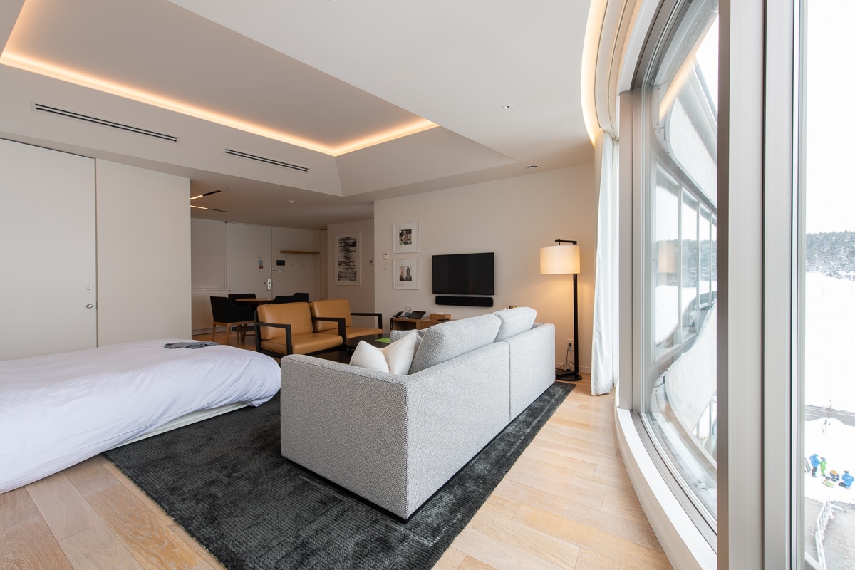 A spacious suite features large windows that provide ample natural light and a view of the surrounding landscape. The seating area includes a light-colored sofa and wooden chairs, while a modern TV is mounted on the wall, enhancing the area.