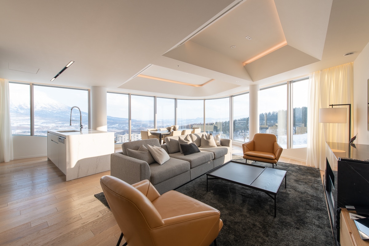 A spacious living area is framed by large windows that illuminate the space, offering views of the surrounding landscape. Comfortable seating is arranged around a central coffee table on a plush area rug. A modern kitchenette is visible in the background, enhancing the open layout.