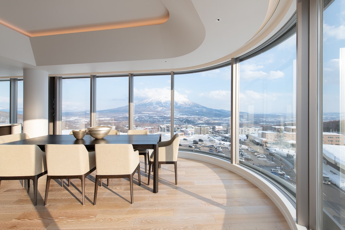 A spacious dining area is surrounded by large curved windows, offering panoramic views of a mountain landscape. A dark wooden table is complemented by light-toned upholstered chairs, and warm natural light enhances the modern design of the space.