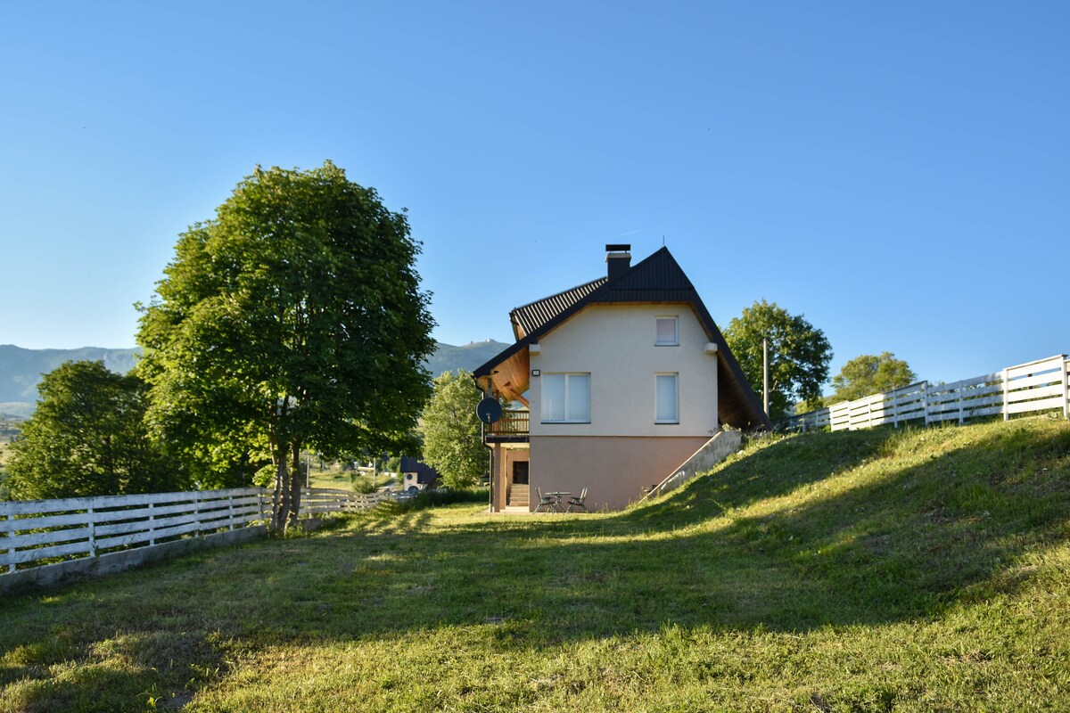 A three-story villa is set amidst a spacious green landscape, with large trees nearby. The exterior is painted in neutral tones, and expansive windows allow for natural light. A white fence outlines the property, enhancing the serene atmosphere of the surrounding area.