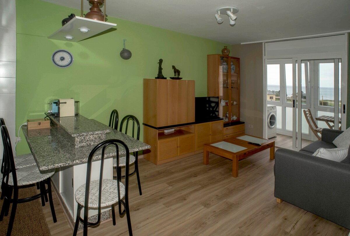 A bright living area features a granite kitchen counter with black chairs, offering a casual dining space. A television is situated on a wooden unit, complemented by glass cabinets. Natural light flows through large windows, revealing distant sea views and enhancing the space's airy ambiance.