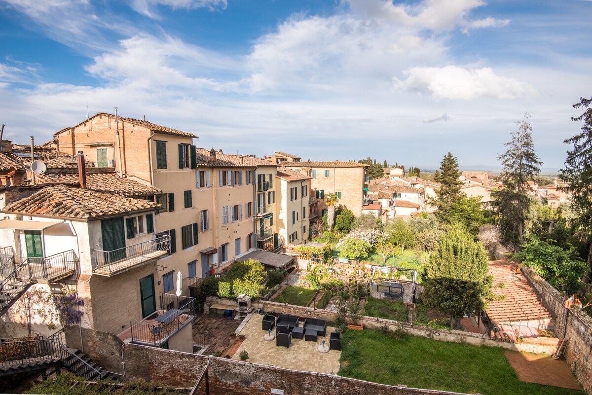 A panoramic view of the surrounding area is captured, featuring rustic buildings with wooden shutters. A garden with various trees and greenery is visible below, alongside a patio area furnished with seating. The sky is bright, with scattered clouds adding depth to the scene.