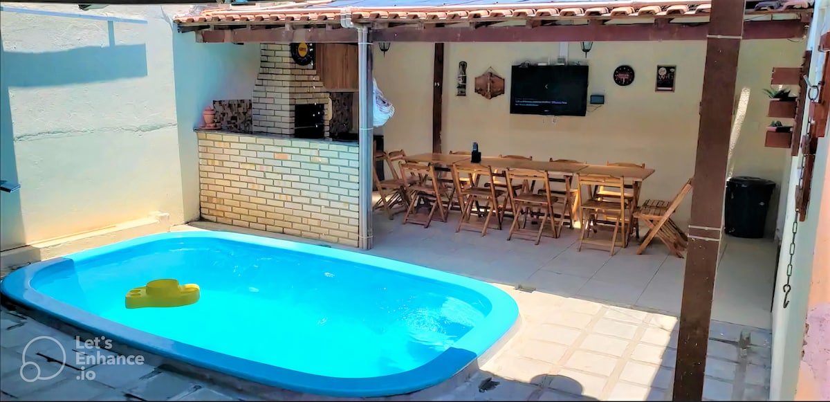 An outdoor area features a swimming pool under a shaded patio with a dining table and chairs arranged for group gatherings. A barbecue grill is situated beside the pool, and a television is mounted on the wall, providing a space for socializing.
