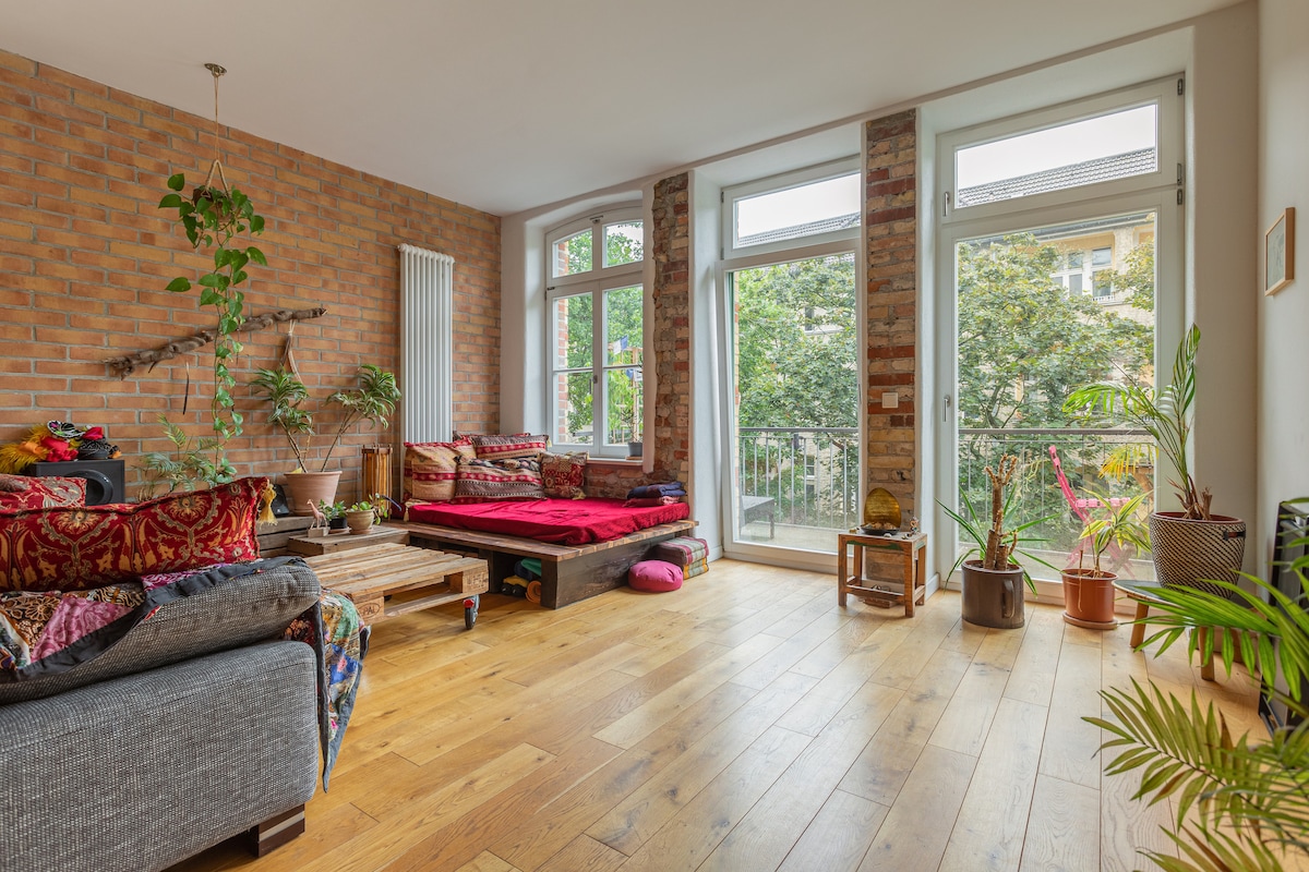 A spacious living area features natural light filtering through large windows that overlook greenery. A cozy seating arrangement, including a low platform bed with vibrant textiles, complements the rustic brick wall. Potted plants add a touch of nature, creating a relaxed environment.