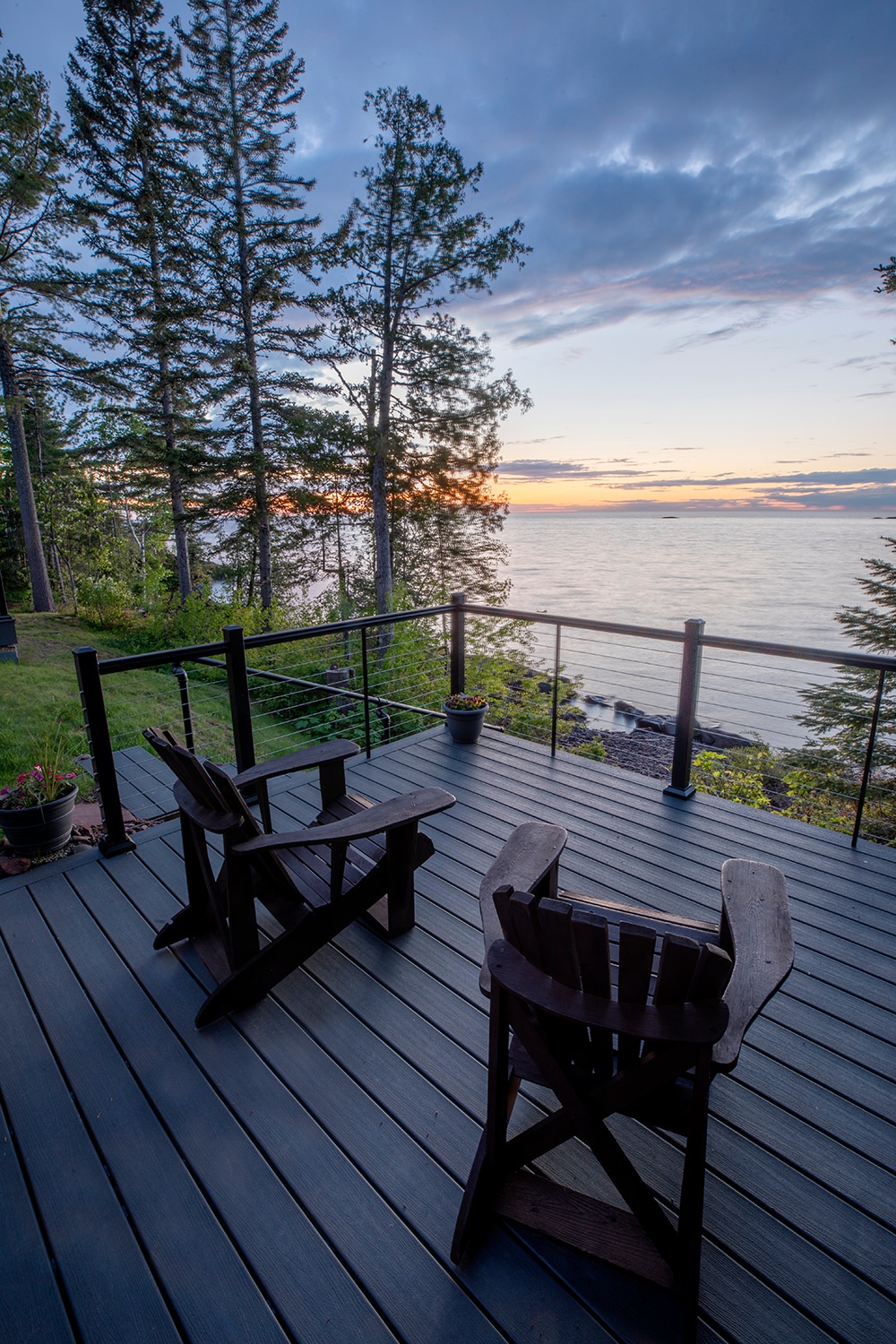 Two wooden chairs are positioned on a spacious deck overlooking Lake Superior. The railing offers unobstructed views of the tranquil water at sunset, with vibrant colors reflecting in the sky. Surrounded by trees, the peaceful environment invites relaxation.