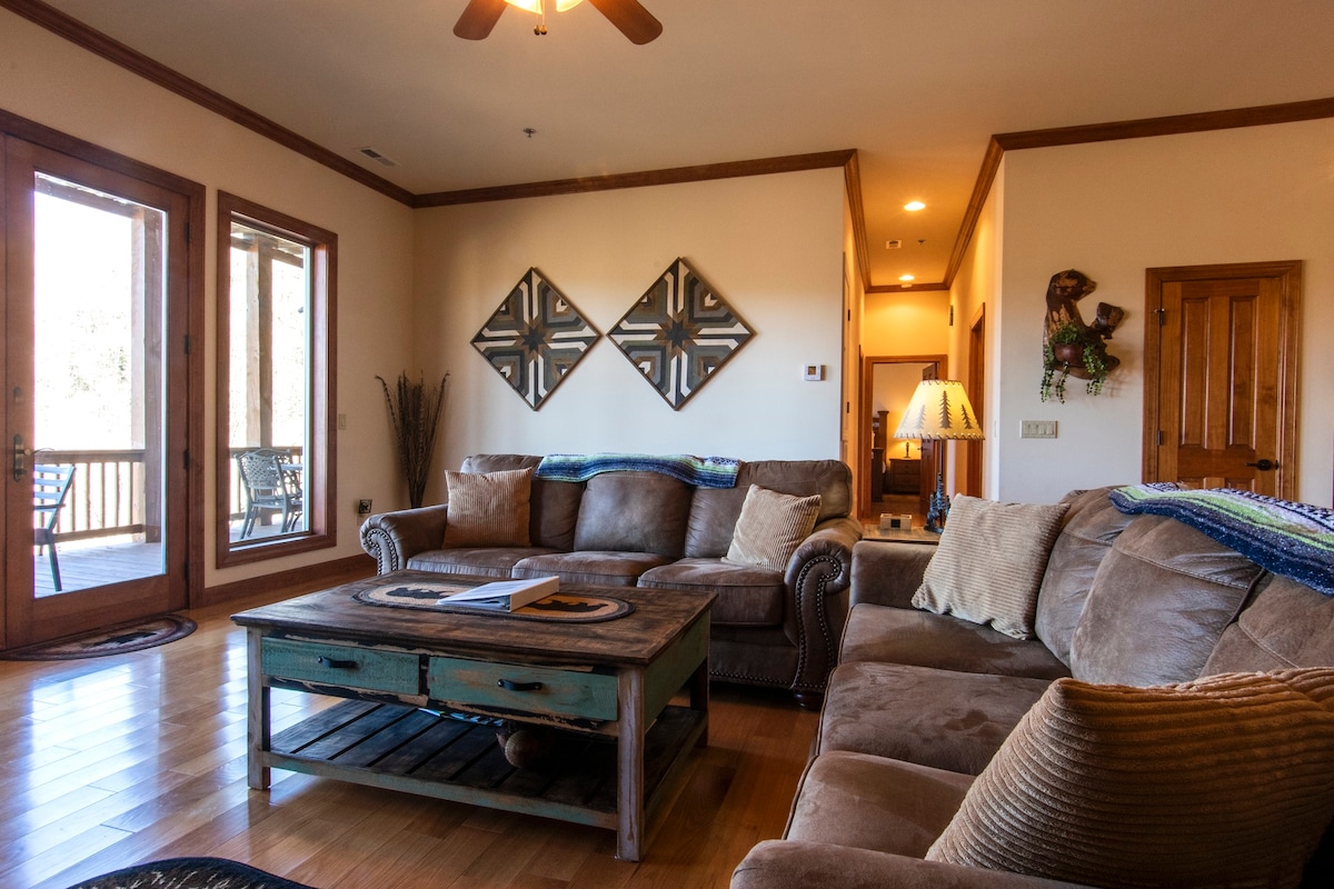 The great room features a comfortable seating area with a large sectional sofa and a rustic coffee table. Light wooden floors and natural light from sliding glass doors enhance the space. Two decorative wall pieces complement the warm decor.
