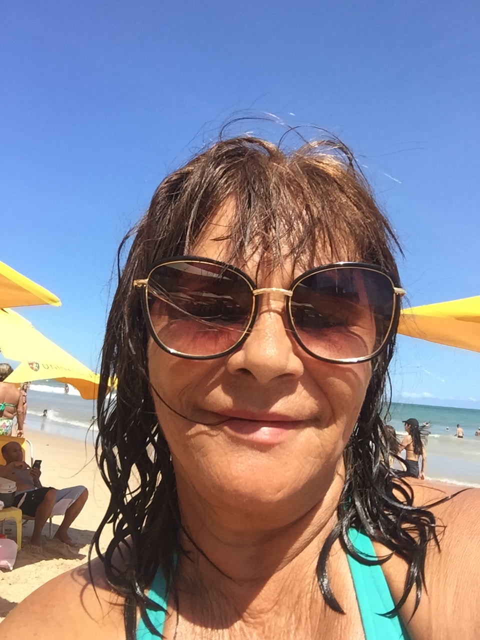 A sunny beach scene features a woman with wet hair in swimwear, wearing sunglasses. Bright yellow umbrellas provide shade in the background, alongside beachgoers enjoying the warm weather and the ocean waves. The clear blue sky complements the vibrant setting.