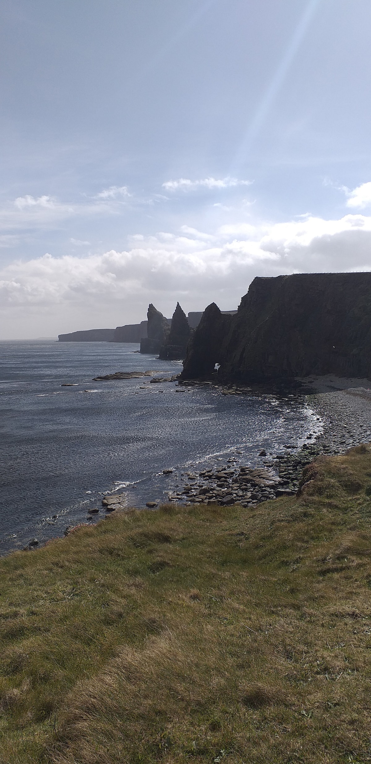 A panoramic view of rugged cliffs meeting the sea, with jagged rock formations rising from the shoreline. Sunlight casts a shimmering effect on the water, while patches of greenery blanket the grassy areas. The coastline extends into the distance under a partly cloudy sky.