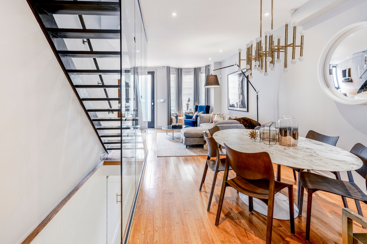 A dining area features a round marble table surrounded by dark wooden chairs, offering a modern aesthetic. The open space is illuminated by a contemporary chandelier. In the background, a cozy living area is visible with a sofa and stylish décor, enhancing the inviting atmosphere.