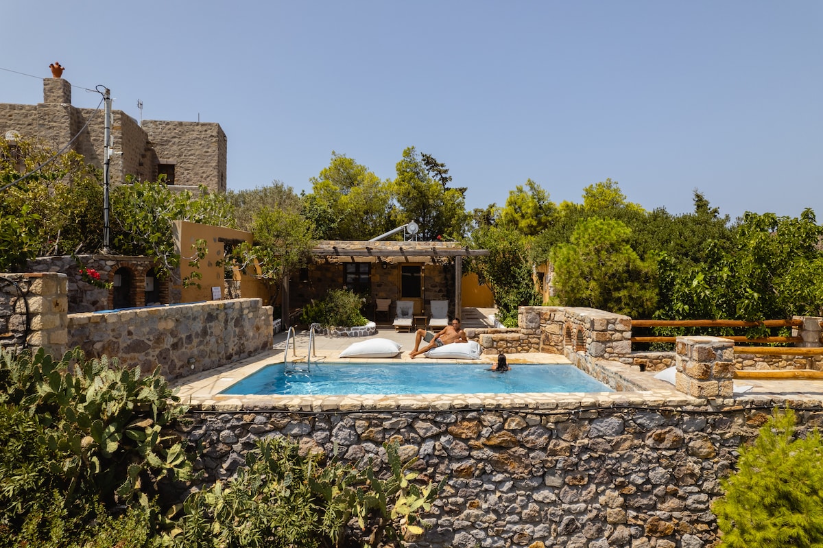A serene outdoor area showcases a private swimming pool surrounded by lush greenery and stone walls. Two loungers are positioned near the pool, inviting relaxation under the sun. The setting reflects a harmonious blend of nature and modern comfort.