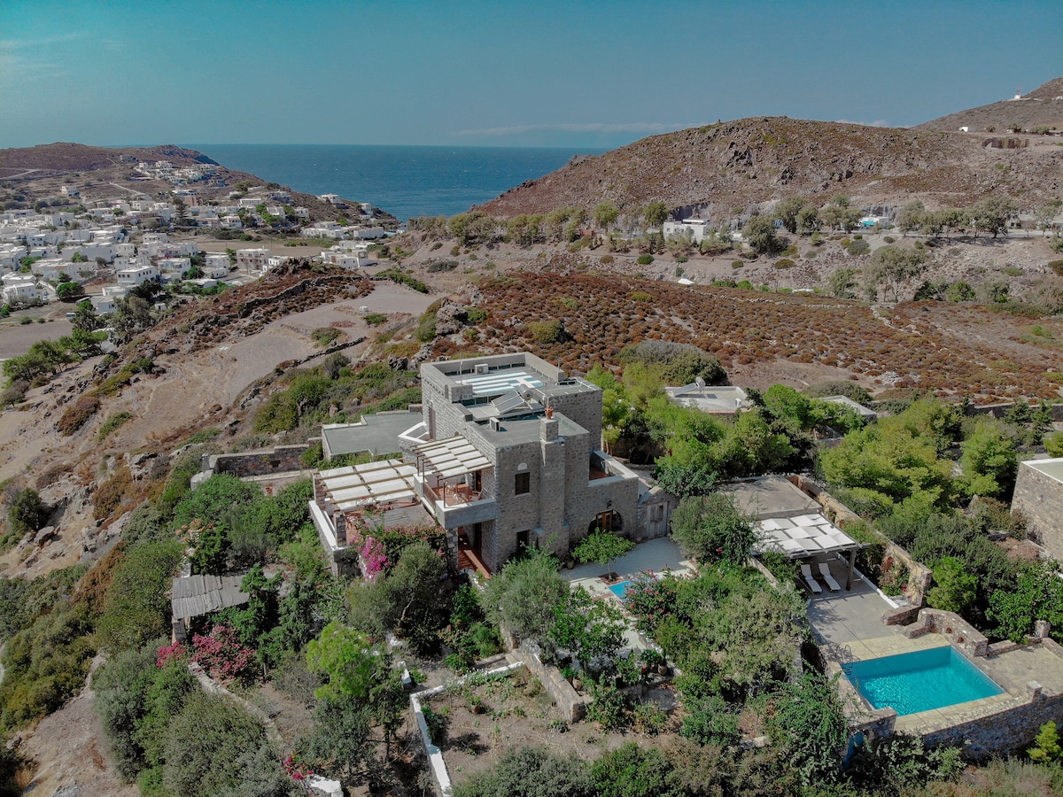 An aerial view of Villa Margarita showcases its stone architecture nestled amidst lush gardens. The villa is surrounded by various trees and flowering plants, with a private swimming pool visible in the foreground. The serene Aegean Sea and hills create a picturesque backdrop.