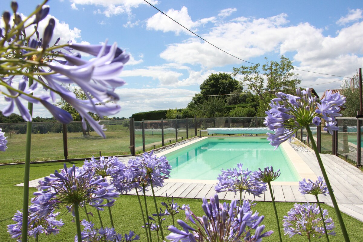 A tranquil swimming pool is surrounded by vibrant flowers, with clear blue water reflecting the sky. The pool area features wooden decking and lush green grass, framed by fencing. In the background, trees and grassy fields add to the serene landscape.
