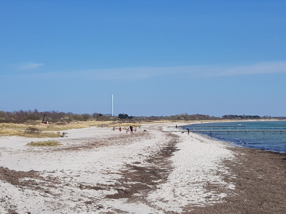 A sandy beach stretches out under a clear blue sky, with gentle waves lapping at the shore. Groups of people can be seen walking along the coastline, with patches of grass and a distant pier visible in the background.