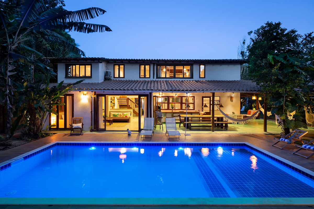 A spacious outdoor area features a well-lit pool surrounded by lounge chairs. The two-story house showcases large glass doors and windows, providing views of the interior. Lush greenery and palm trees enhance the tropical ambiance, creating a serene setting for relaxation.