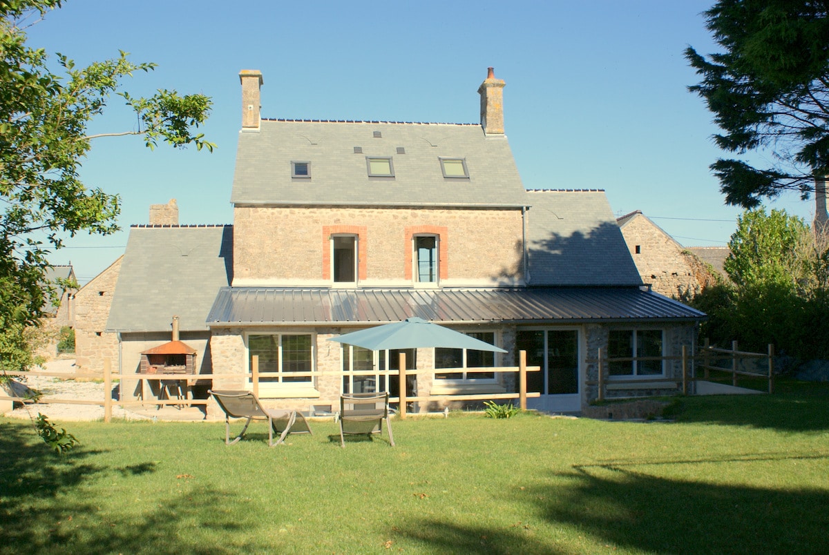 The exterior of the restored farmhouse presents a blend of stone and slate, framed by a lush green lawn. Sun loungers are positioned on the grass, while a blue umbrella provides shade. A barbecue area is visible nearby, enhancing outdoor dining possibilities.