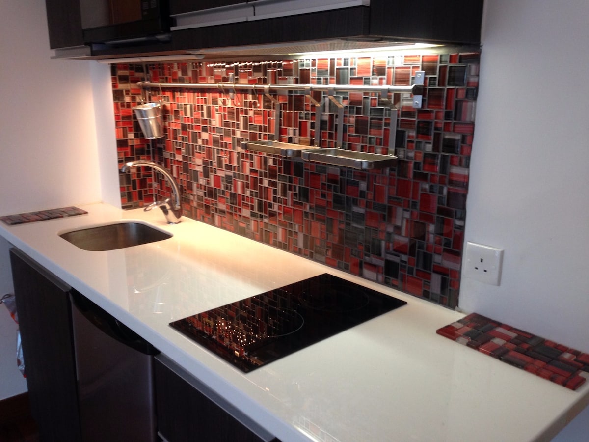 A modern kitchen area features sleek cabinetry and a vibrant mosaic backsplash in shades of red and gray. Counter space is complemented by a stainless steel sink, and an induction cooktop is seamlessly integrated into the counter design.