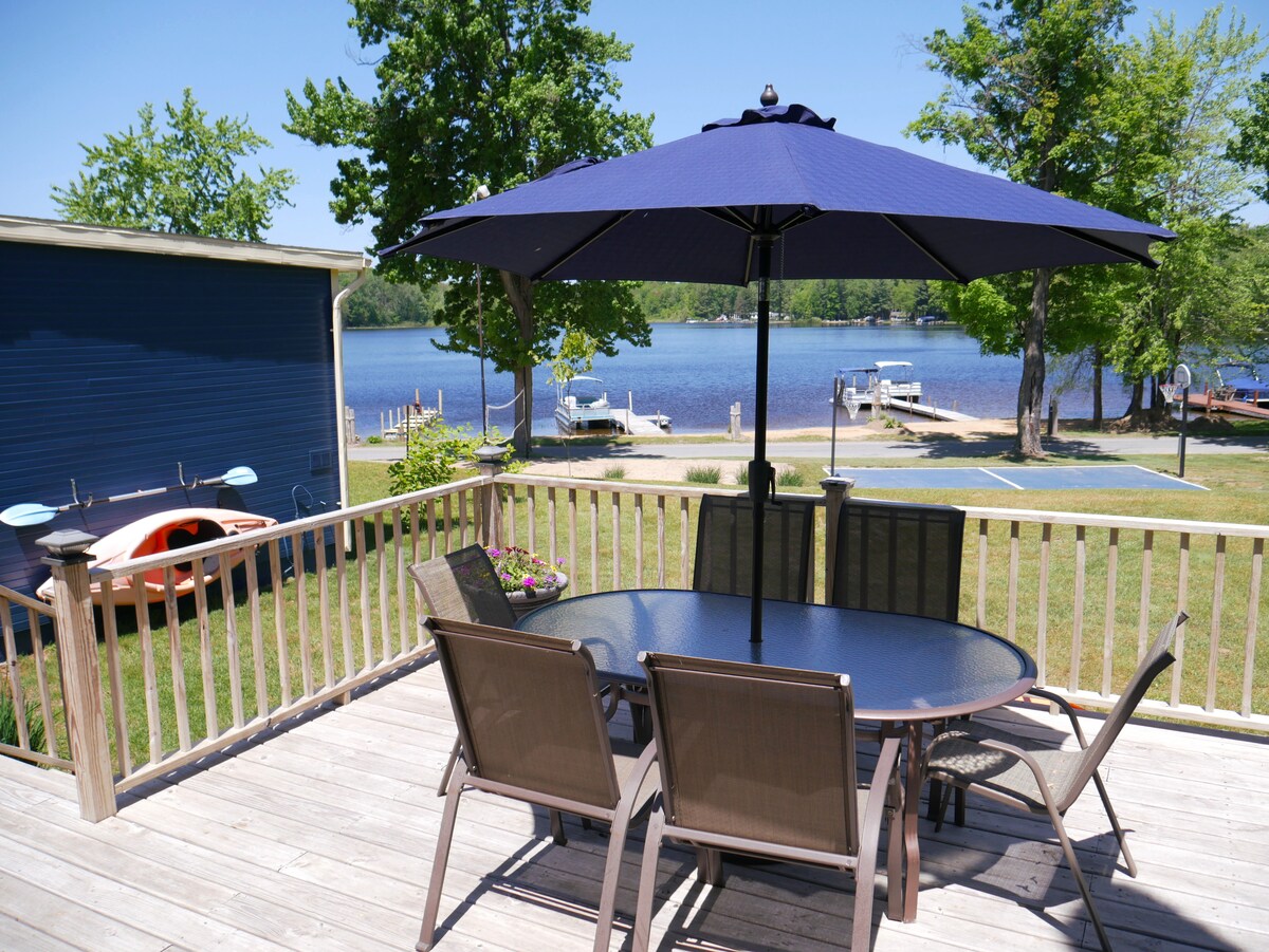 A shaded outdoor dining area features a round table and six chairs under a large blue umbrella. The deck overlooks a serene lake with boat docks visible in the distance, surrounded by lush greenery and clear blue skies.
