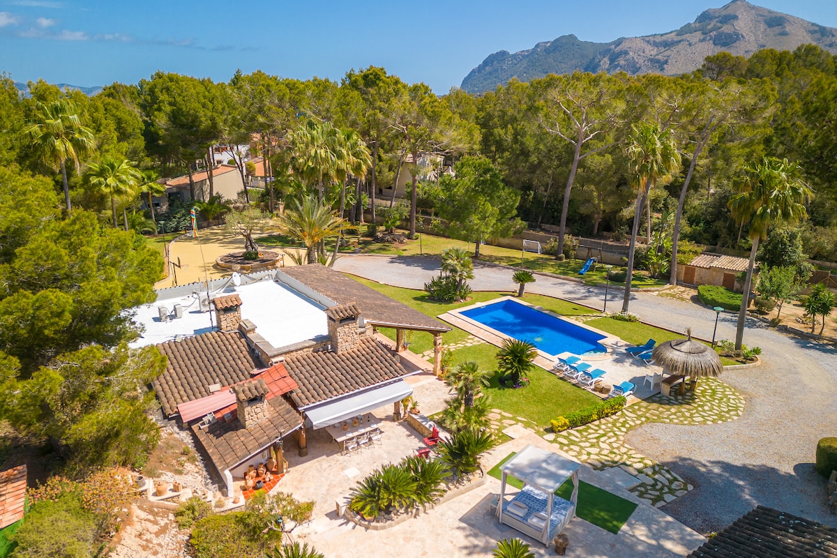 An aerial view of a spacious country house surrounded by greenery and palm trees. A private swimming pool is visible alongside a sun patio, offering ample space for relaxation. The landscape features pathways and various outdoor areas, enhancing the serene ambiance of the property.