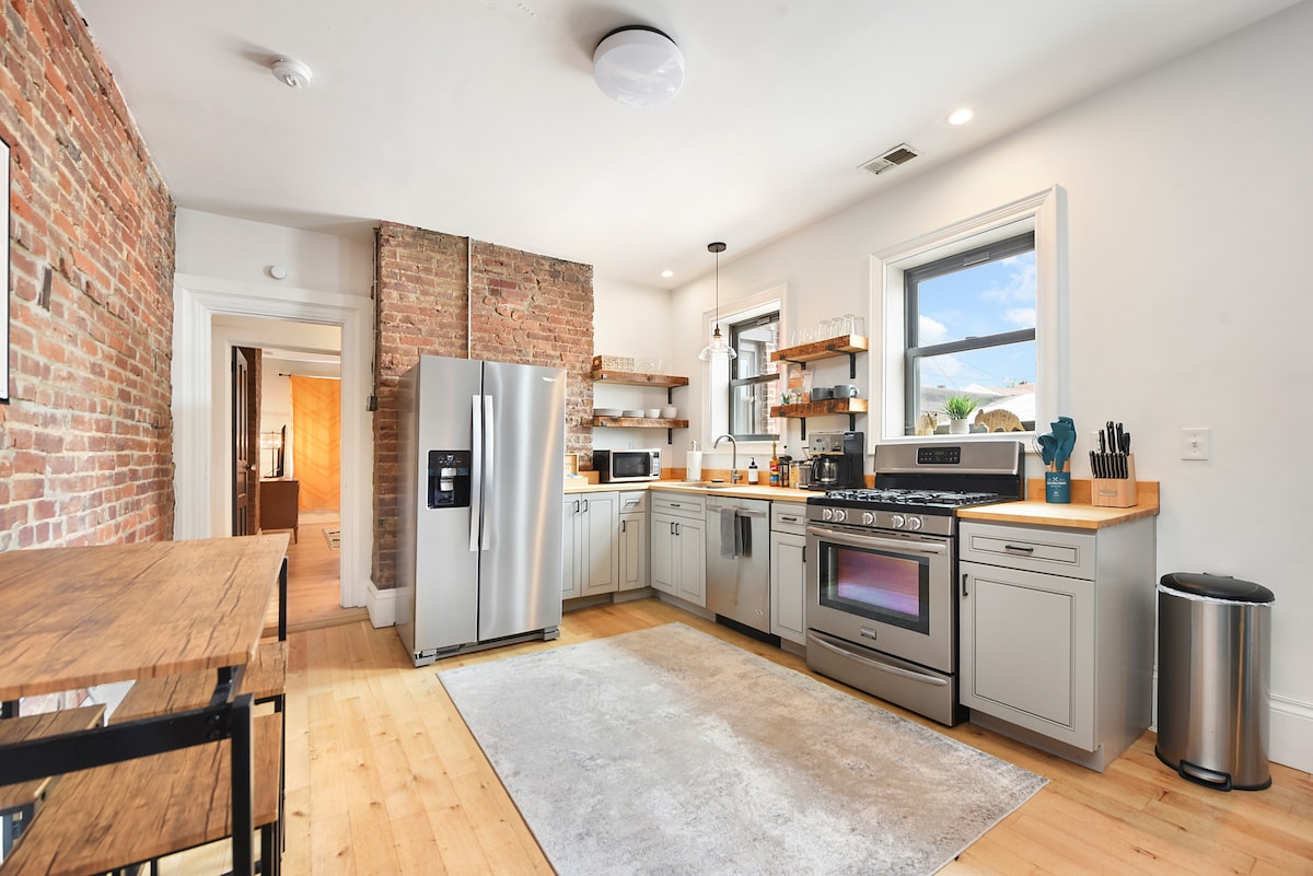 A modern kitchen features stainless steel appliances, including a large refrigerator and a sleek stove with an oven. The minimalist cabinetry is complemented by exposed brick walls. Natural light streams through the windows, illuminating the wood countertops and open shelving.