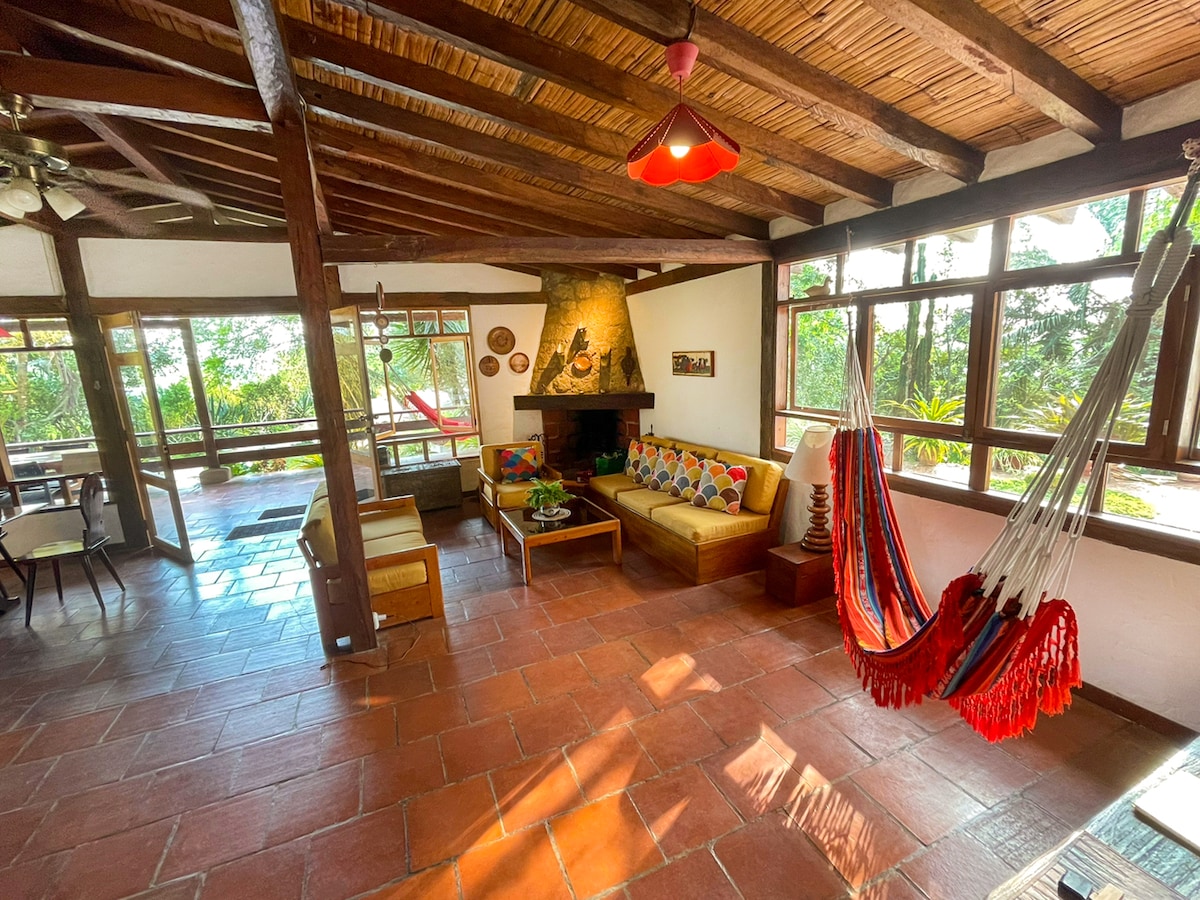 A spacious living area features comfortable seating, a central stone fireplace, and large windows that allow natural light. Vibrant cushions adorn the seating, while a colorful hammock hangs nearby. The terracotta floor and wooden beams enhance a warm atmosphere in this inviting space.