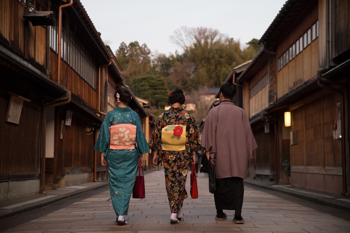 Three individuals dressed in traditional Japanese kimonos are walking along a wooden pathway between historic buildings. The scene captures their attire, featuring a blend of patterns and colors. The soft evening light enhances the serene ambiance of the surrounding area.