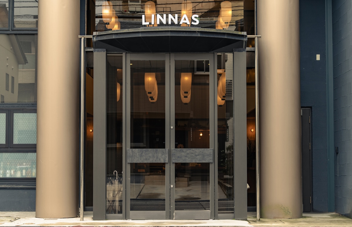 The exterior entrance of LINNAS Kanazawa is showcased with a large glass door framed by beige columns. The name 'LINNAS' is prominently displayed above. Soft lighting from inside creates a welcoming ambiance visible through the glass.