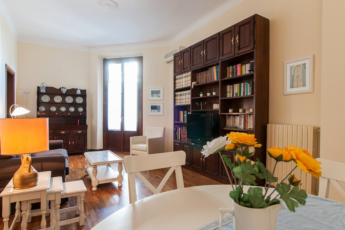 A living area is presented with hardwood flooring and neutral-colored walls. Shelving units filled with books and decorative items provide visual interest. A cozy seating arrangement includes a sofa, armchair, and a coffee table. A vase of flowers sits on the dining table, enhancing the space.