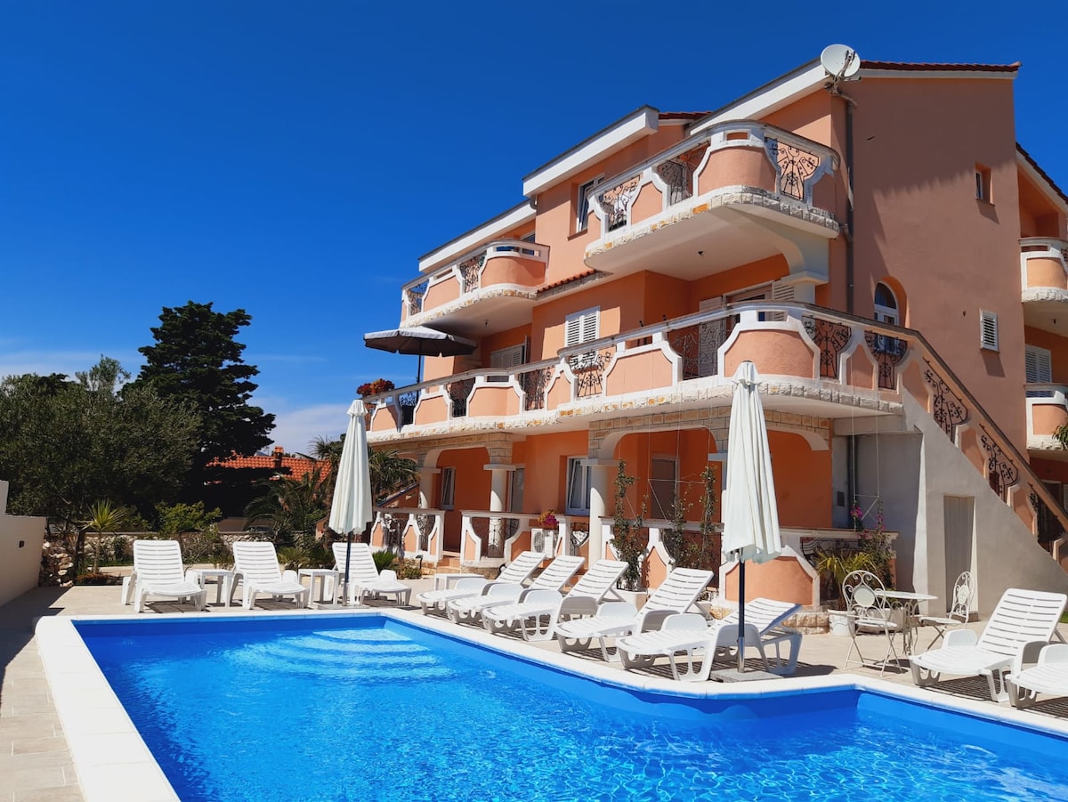 A well-maintained outdoor area features a bright blue swimming pool surrounded by poolside loungers. A multi-story building with orange stucco walls and decorative balconies is positioned in the background, complemented by clear blue skies and greenery.