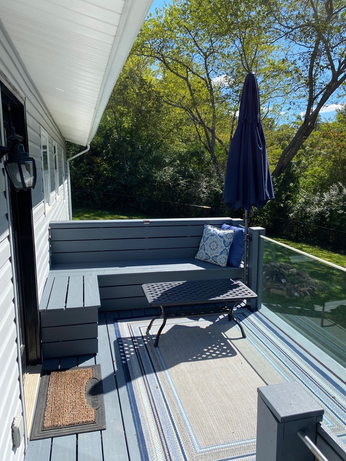 An outdoor seating area is presented with a wooden bench adorned with a decorative cushion. A small, black, metal table sits on a patterned rug beneath a large umbrella. Lush greenery is visible in the background, providing a serene setting.