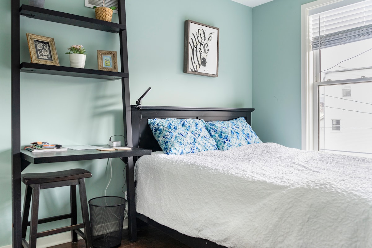 A cozy room is presented with a queen-size bed dressed in a white quilt and blue-patterned pillows. A small desk and chair are positioned beside the bed, with a light fixture and a wastebasket nearby. Light enters through a window adorned with blinds, complementing the calming wall color.