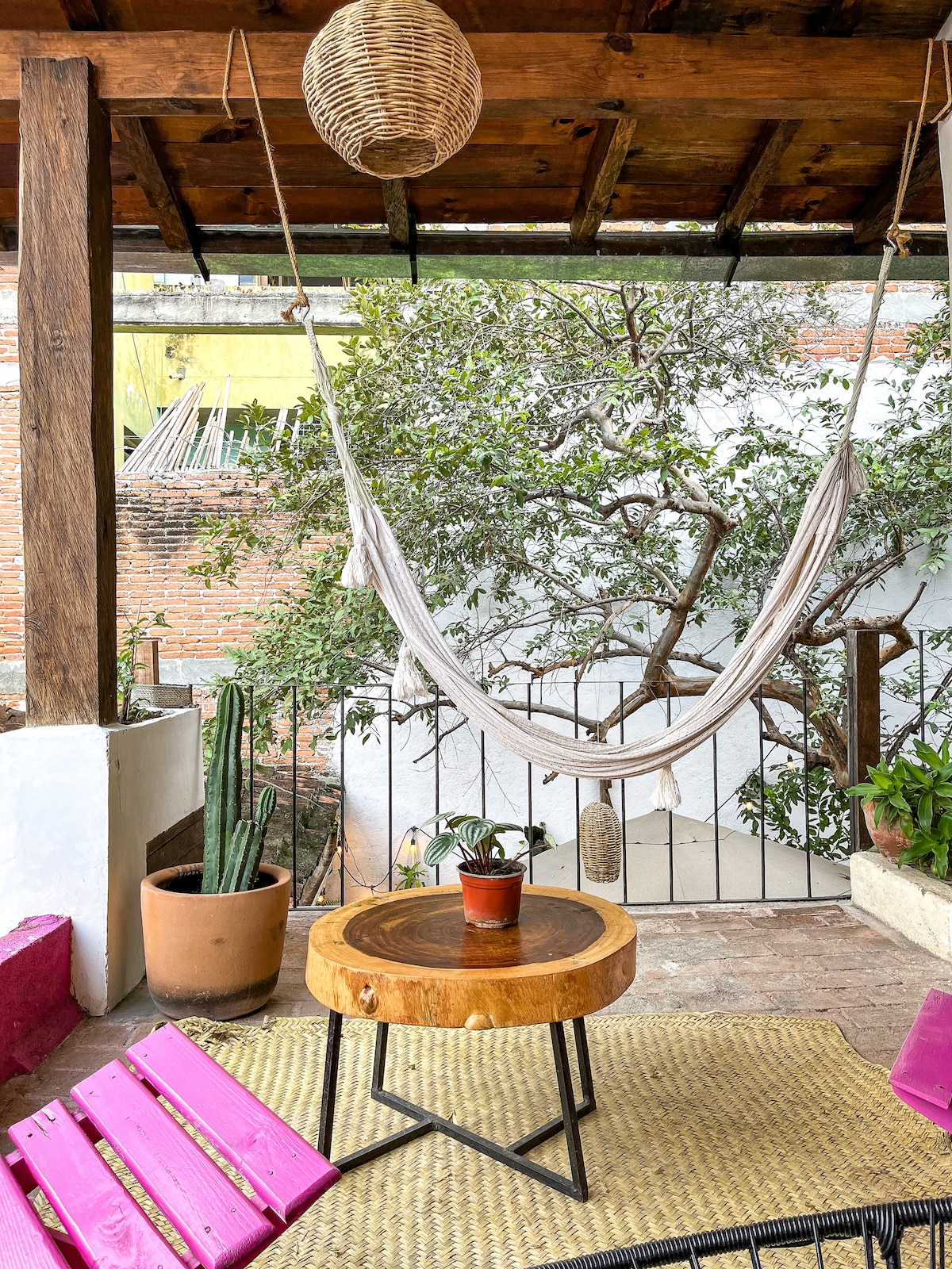 A relaxing outdoor area features a hanging hammock draped between wooden beams. A circular wooden table with a small potted plant is positioned nearby, with pink benches lining the space. Green plants and a tall cactus add to the refreshing ambiance.