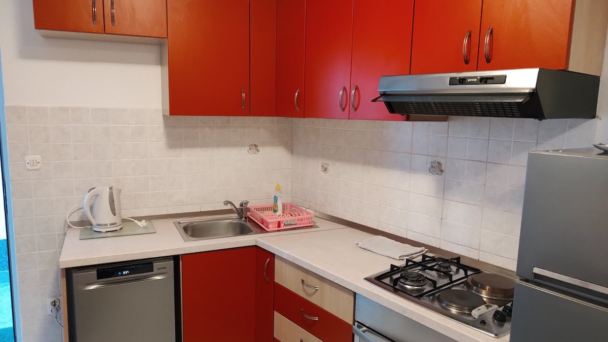 The kitchen is equipped with modern appliances, including a stove, refrigerator, and dishwasher. Red cabinetry provides a vibrant contrast against the light tiled walls. A sink and kettle are also present, along with a pink dish drying rack on the countertop.