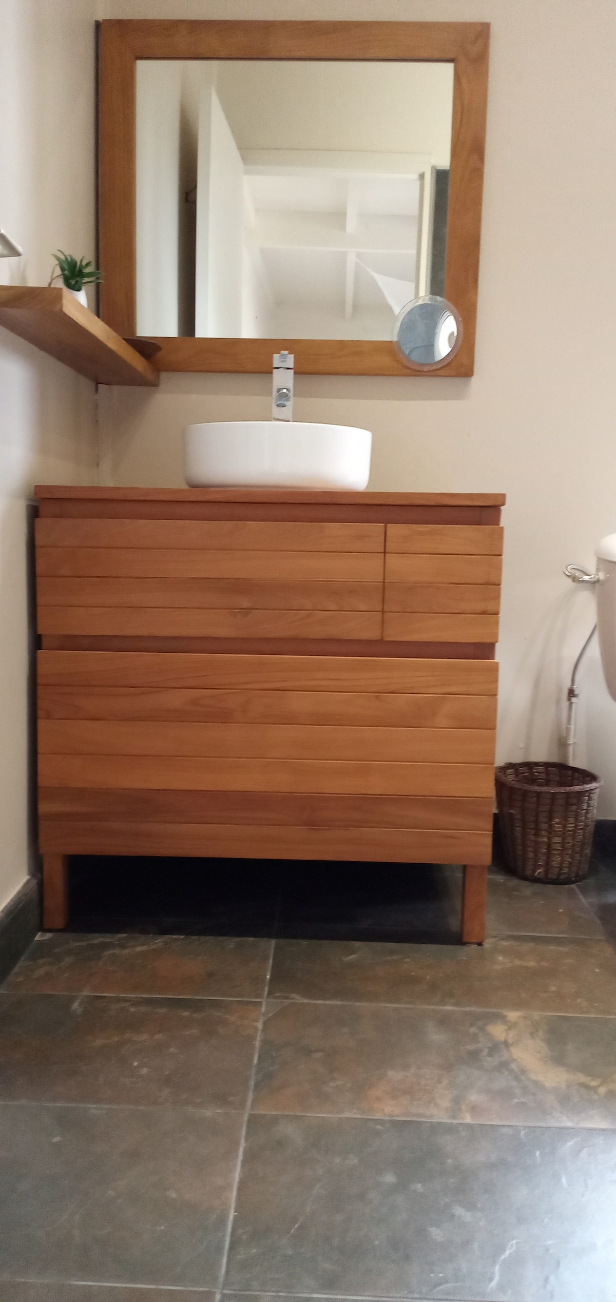 A wooden vanity unit features a minimalist design with two drawers and open space below. A round basin is positioned atop the surface, reflecting the natural light from a nearby window. A framed mirror hangs above, offering a functional addition to the space.