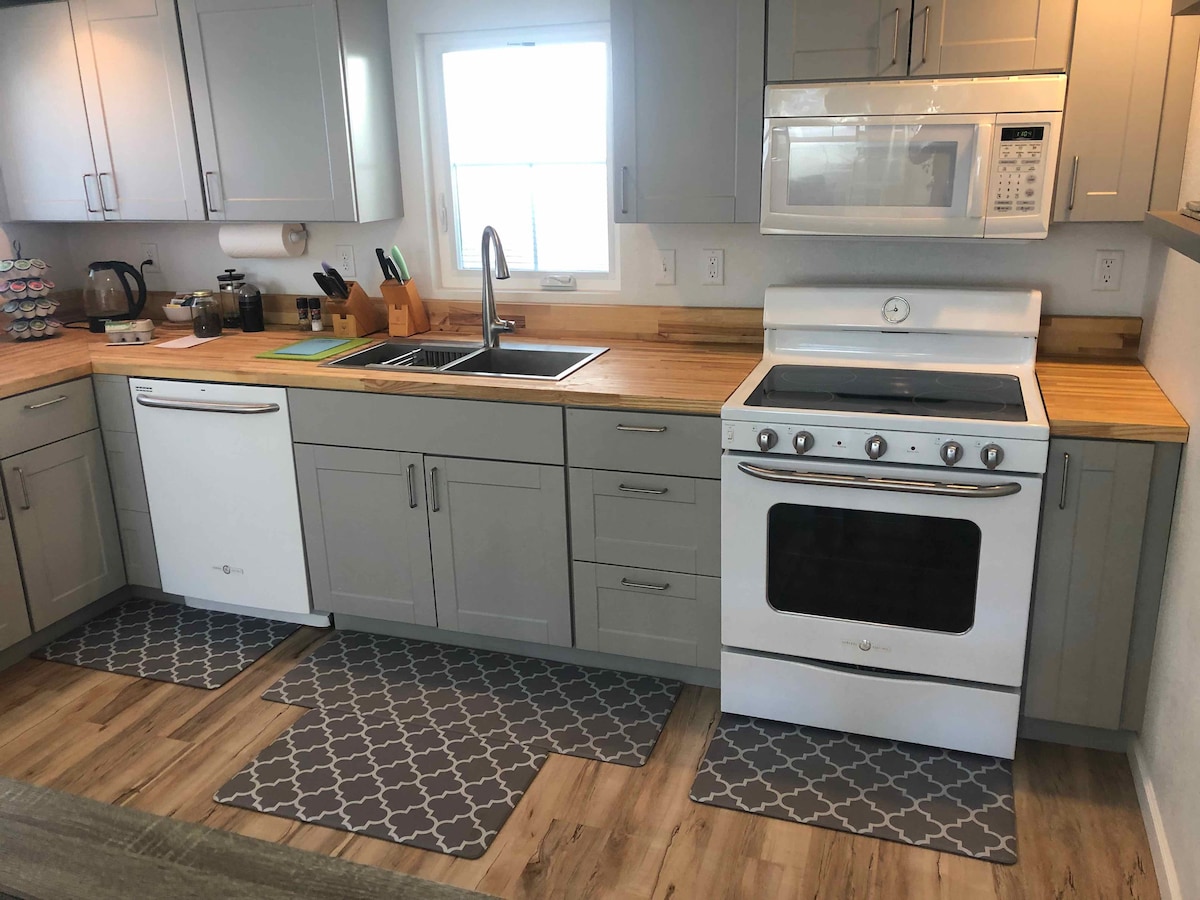 A modern kitchen is presented with gray cabinetry and light wooden countertops. A gas stove and microwave are seen alongside a double basin sink. Kitchen mats in a geometric pattern are placed on the floor, contributing to the functional layout.