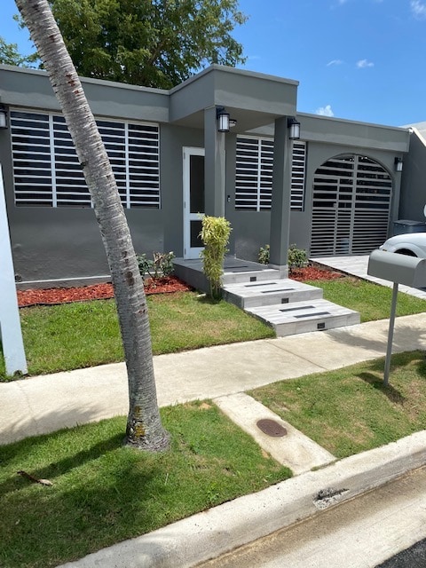 The exterior of the home features a modern design with a gray façade. A walkway of steps leads to the front door, surrounded by neatly landscaped grass and tropical plants. The façade includes large windows with horizontal slats, providing natural light while maintaining privacy.