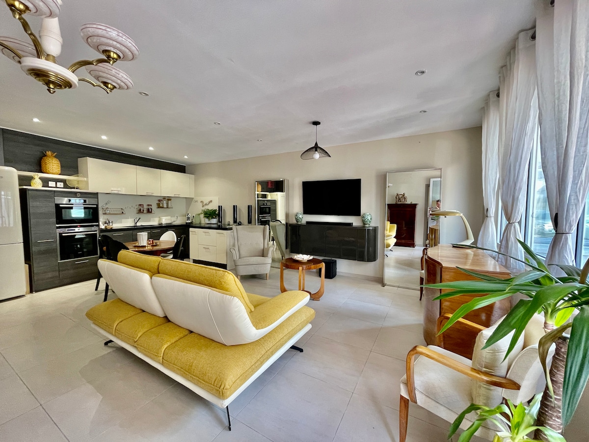A spacious living area is outlined by large windows, allowing natural light to fill the room. A modern yellow and white sofa complements a wooden accent table and elegant armchair. The kitchen is visible, featuring sleek cabinetry and high-end appliances.